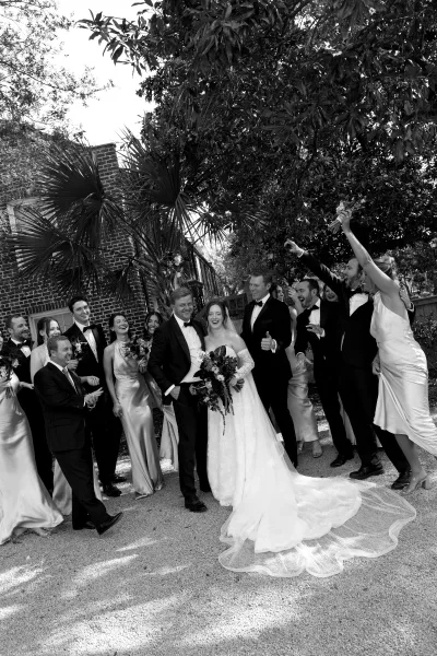 Wedding party portrait captures a candid wedding party cheering, bride holding a lush bridal bouquet before a brick wall framed by palm leaves.