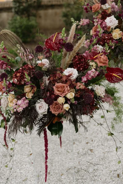 Wedding floral arrangement of ceremony flowers in burgundy and blush on a clear acrylic stand, set in a garden by a stone wall.