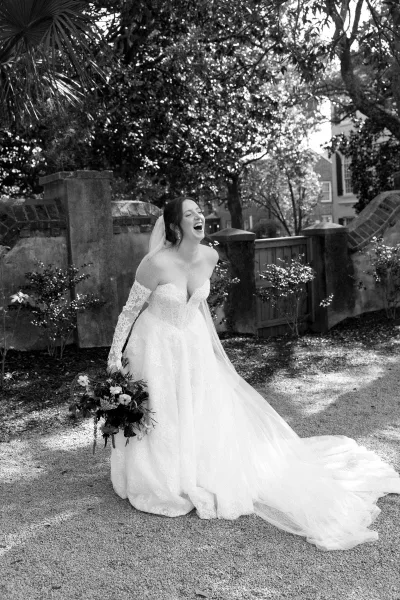 Bridal portrait of a laughing bride holding a bridal bouquet, framed by garden trees beside a weathered stone wall and wooden gate.