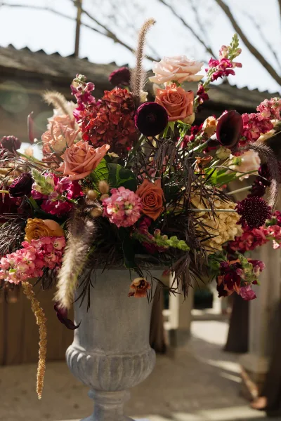 Wedding floral arrangement styled as an urn floral arrangement with dried grasses, set in a sunlit outdoor courtyard framed by stone columns.