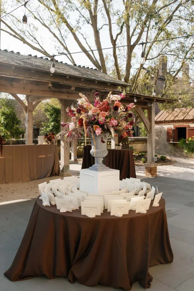 Escort card display on an escort card table topped with a floral arrangement, under a wooden pavilion overlooking trees and a stone patio.