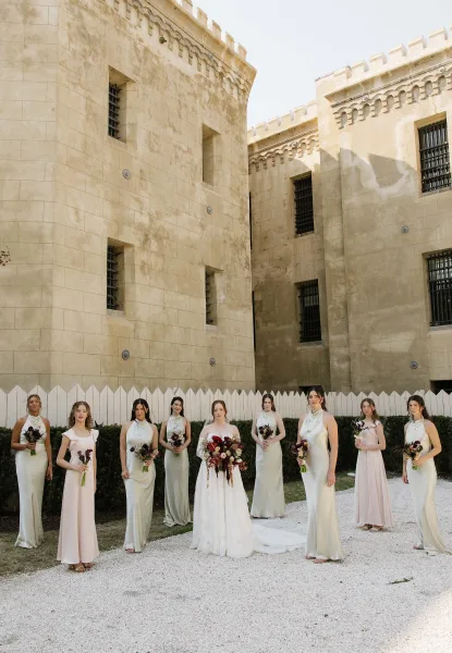Bride and bridesmaids pose in a lineup, inspired by bridesmaid photo ideas, the bridal bouquet framed by a stone building and white picket fence.