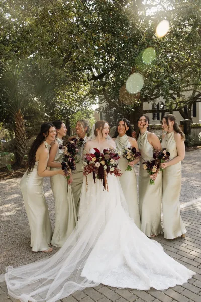 Bride and bridesmaids laugh in an outdoor bridal party photo on a sunlit garden walkway, the bride’s cathedral veil flowing behind her.