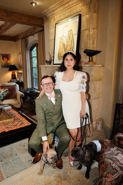 Couple portrait of bride and groom with dogs, posed by a stone fireplace with framed artwork in a cozy living room.