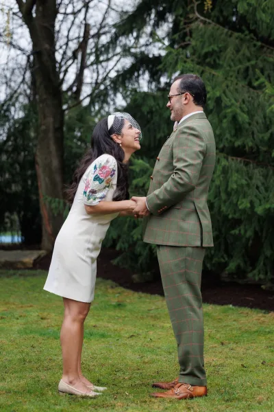 Couple portrait captured as a garden wedding portrait, the bride in a birdcage veil laughing as they hold hands among evergreen trees.
