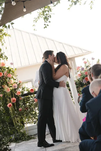 Ceremony kiss moment as the bride and groom share their wedding first kiss beneath string lights, framed by a white roof and greenery vines.