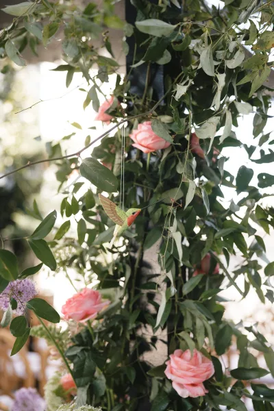 Wedding floral decor with a greenery and roses installation, a hummingbird ornament, and garden foliage framing nearby wooden chairs.