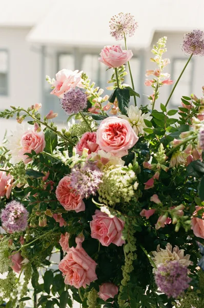 Wedding floral arrangement with pink garden roses and trailing greenery, styled in soft daylight against a white building with tall windows.