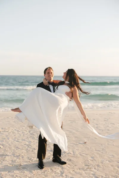 Couple portrait with the groom carrying the bride, his bow tie visible, on a sandy beach by rolling ocean waves.