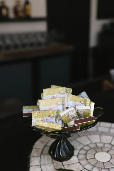 Matchbox wedding favors and wedding matchbooks styled in a pedestal bowl on a tiled bar, with glassware and liquor bottles behind.