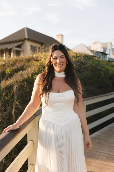 Bridal portrait of a bride on a boardwalk, smiling in a strapless satin gown with delicate earrings, houses and shrubbery softly behind.