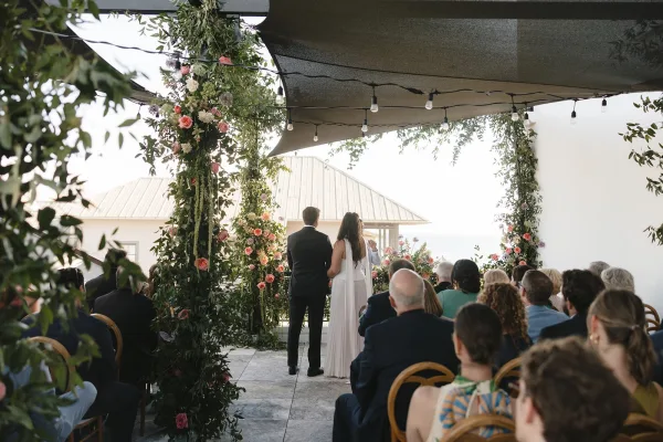 Ceremony moment with the couple at the altar beneath a floral arch, exchanging vows on a canopy-shaded patio with an ocean view.