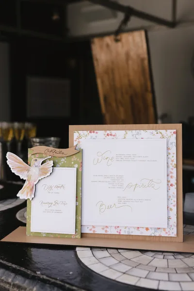 Wedding bar sign and cocktail menu sign with a die-cut hummingbird detail, propped on a tiled tabletop.