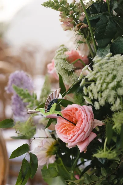 Floral arrangement styled as a garden rose centerpiece, highlighted by a delicate hummingbird ornament for a whimsical, romantic touch.