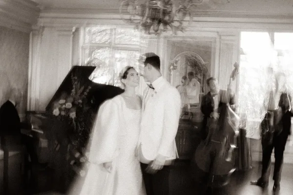 Couple portrait of a bride and groom with band, smiling beside a grand piano as musicians frame the moment and the newlyweds share a warm gaze.