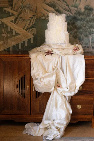 Wedding cake, a white tiered cake styled with draped fabric, displayed on a wooden cabinet against mural wallpaper and hardwood floor.