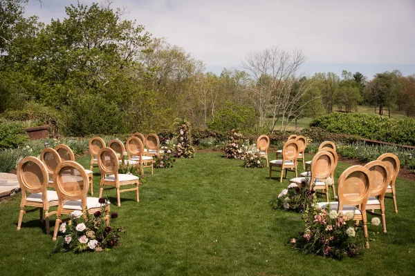 Outdoor ceremony setup with elegant garden ceremony decor, natural wood chairs arranged on a grass lawn beneath garden trees and hedges.