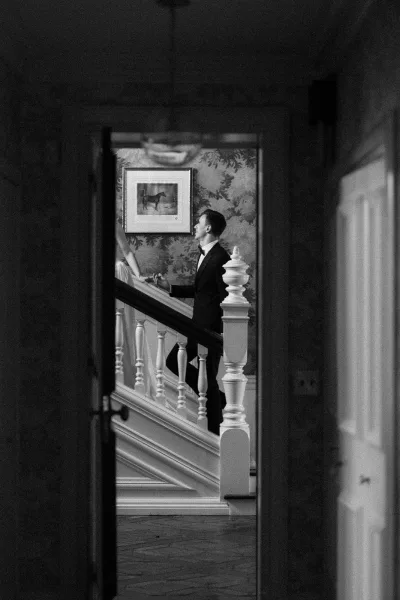 Couple portrait of a bride and groom on stairs, the groom in a tuxedo, framed by a doorway, with wallpapered wall and framed artwork in view.