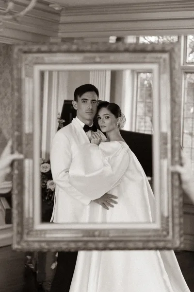 Couple portrait as a black and white wedding portrait, framed by a vintage picture frame as they embrace beside a grand piano in a stately room.