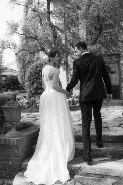 Couple portrait of the bride and groom walking hand in hand, her hair flower catching light, on stone steps by a brick building and garden trees.