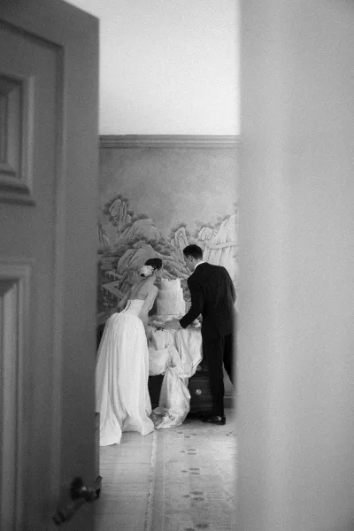 Wedding cake cutting as the bride and groom with cake stand together in a quiet moment, his black suit framed against a mural wall.