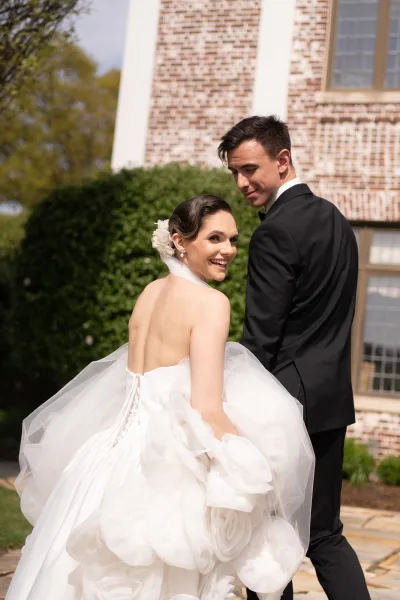 Couple portrait of newlyweds walking away, the bride lifting a ruffled tulle skirt as they stroll down a stone path beside a brick wall and hedge.