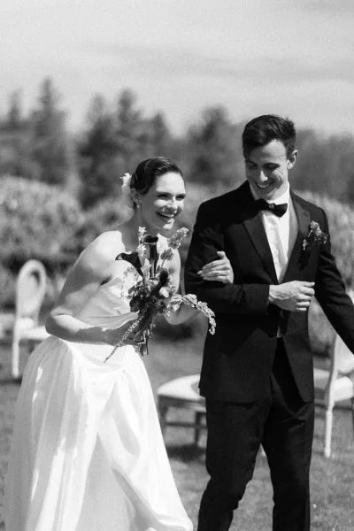 Ceremony exit with the couple walking down the aisle, bride and groom laughing as she holds a bouquet, garden trees and chairs on the lawn.