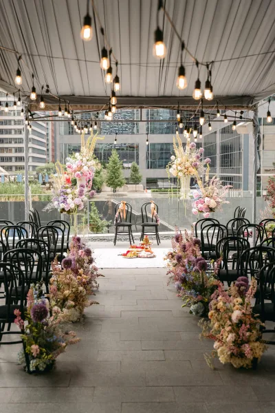 Ceremony setup with outdoor wedding ceremony aisle flowers, black chairs and layered rugs under string lights on a terrace with glass railing