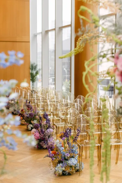 Ceremony aisle decor with clear acrylic ceremony chairs and floral-lined greenery, set in an indoor venue with tall windows and wood walls