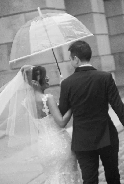 Couple portrait of bride and groom walking under a clear umbrella, her lace dress and veil flowing as she holds a bouquet by a stone wall
