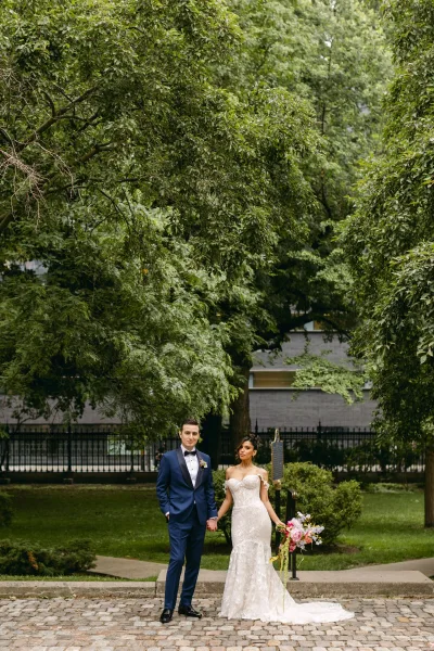 Couple portrait of bride and groom holding hands, her strapless lace wedding dress with train and bouquet on a cobblestone path in a park