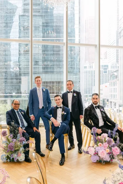 Groomsmen portrait with men in tuxedos posing by gold chairs, wearing boutonnieres, against floor-to-ceiling windows and city skyline view