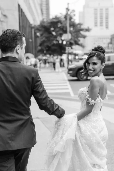 Couple portrait in black and white, bride and groom walking hand in hand at a crosswalk, her lace gown train and veil flowing on a city street
