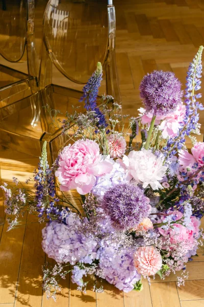 Wedding floral arrangement floor flower arrangement with peonies, allium, hydrangea, lupine and dahlia beside acrylic chairs on sunlit wood floor
