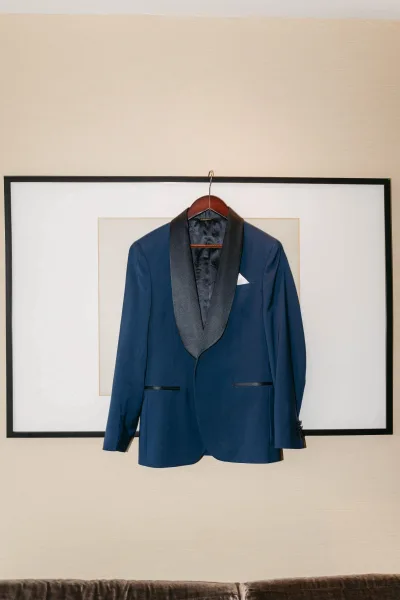 Groom suit detail with a navy tuxedo jacket on a wooden hanger, satin shawl lapel and pocket square against a beige wall with framed art