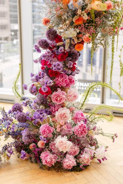 Wedding floral installation with an asymmetrical floral arch of dahlias, roses, and hanging greenery before large windows and city buildings