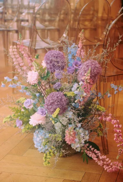 Wedding floral arrangement of pastel hydrangea, allium, and lupine with greenery beside acrylic chairs on an indoor wood floor aisle