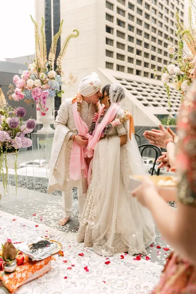 Wedding kiss during a hindu wedding ceremony as the Indian bride and groom wear garlands on a city rooftop terrace with guests clapping