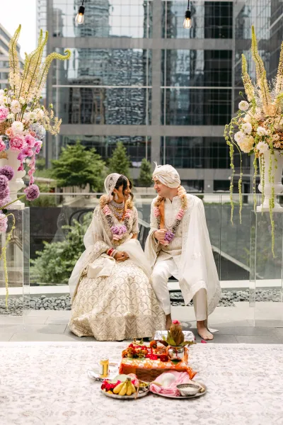 Couple portrait of an Indian wedding couple laughing with floral garlands on a terrace patio, city skyline behind glass railing