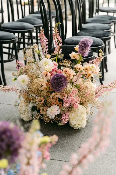 Ceremony aisle florals with pastel pink roses and purple allium arranged low beside black chairs on a stone-floored indoor aisle