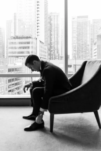 Groom portrait in a black tuxedo seated in a lounge chair, tying dress shoes by a floor-to-ceiling window with city skyline view