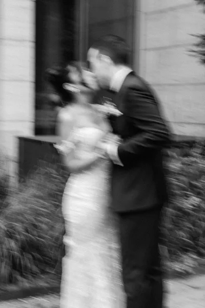 Wedding kiss in a black and white wedding photo, bride in strapless dress holding bouquet as groom in tuxedo leans in by a stone doorway