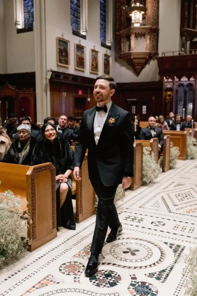 Groom processional as groom walking down aisle in a black tuxedo with boutonniere, candlelit church aisle and stained glass windows behind