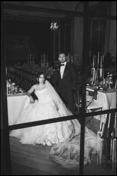 Couple portrait in a black and white wedding portrait, bride seated in strapless gown and cathedral veil as groom stands in tuxedo amid chandeliers and candlelit tables