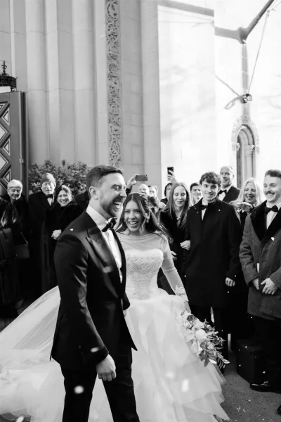 Wedding recessional as newlyweds walk out of church, bride in long veil holding bouquet beside groom in tux, guests filming by stone columns