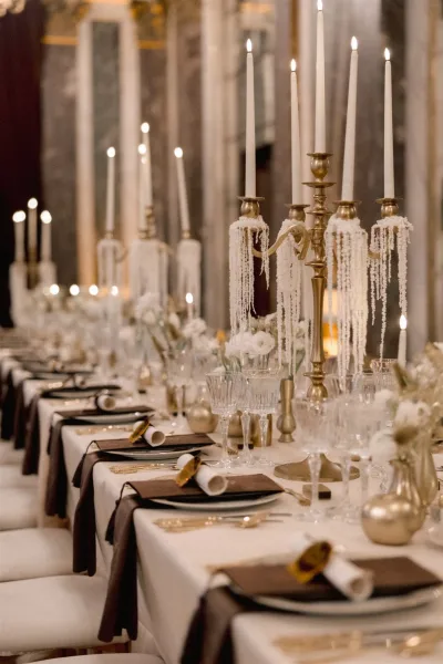 Reception tablescape on a long banquet table with gold candelabras, taper candles, white florals and crystal glassware beneath arched windows