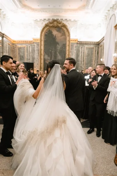 Reception entrance as newlyweds grand entrance through a ballroom aisle, bride in cathedral veil and lace train beside groom in tuxedo, guests clapping