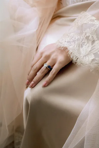Bridal ring close-up showing wedding ring details on a hand with gold pear-shaped and blue gemstone rings, lace sleeve and tulle veil on neutral fabric