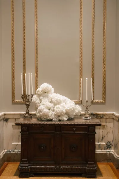 Wedding floral arrangement with a white hydrangea centerpiece, candelabras and taper candles on an antique console against marble wainscoting