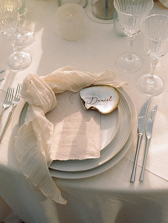 Wedding place setting with wedding menu card on layered plates, oyster shell place card, chiffon napkin and crystal glasses in soft sunlight
